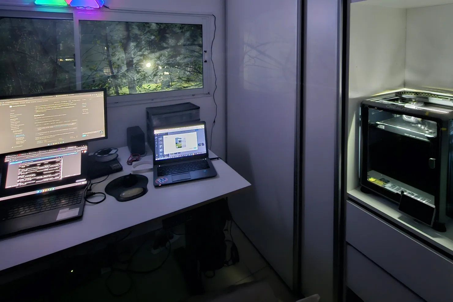 Rincón del home lab de noche: un escritorio con dos notebooks y un monitor mostrando código, paneles de luz RGB triangulares en la pared y una impresora 3D en el mueble contiguo.