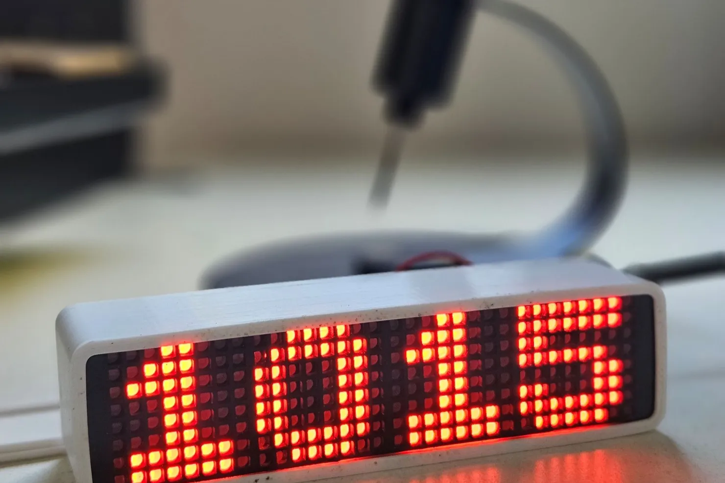 Reloj de matrices LED rojas en una carcasa impresa en 3D blanca, apoyado sobre un escritorio, mostrando la hora en dígitos de puntos.